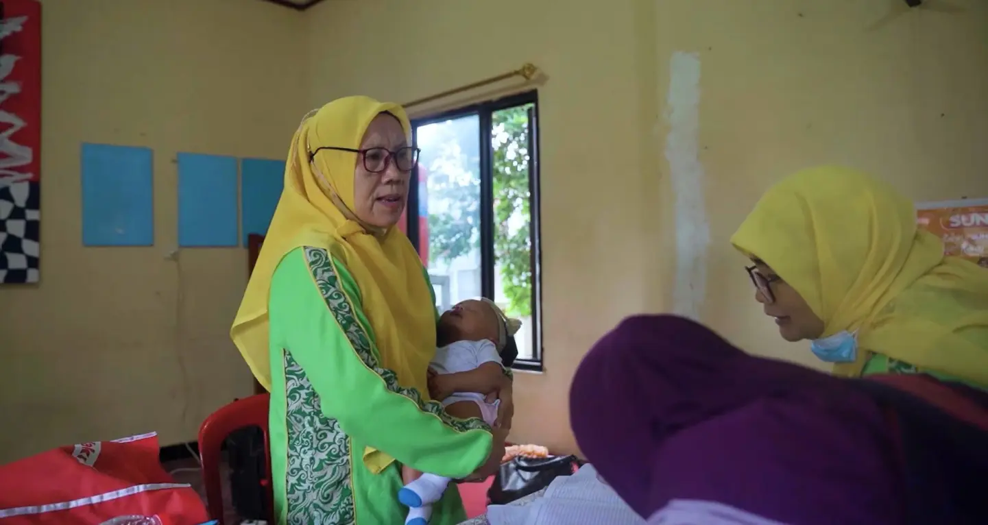 A community health worker wearing a yellow veil carries a baby doll inside a Posyandu room. In front of her, two other CHWs are engaged in the Posyandu educational activities.